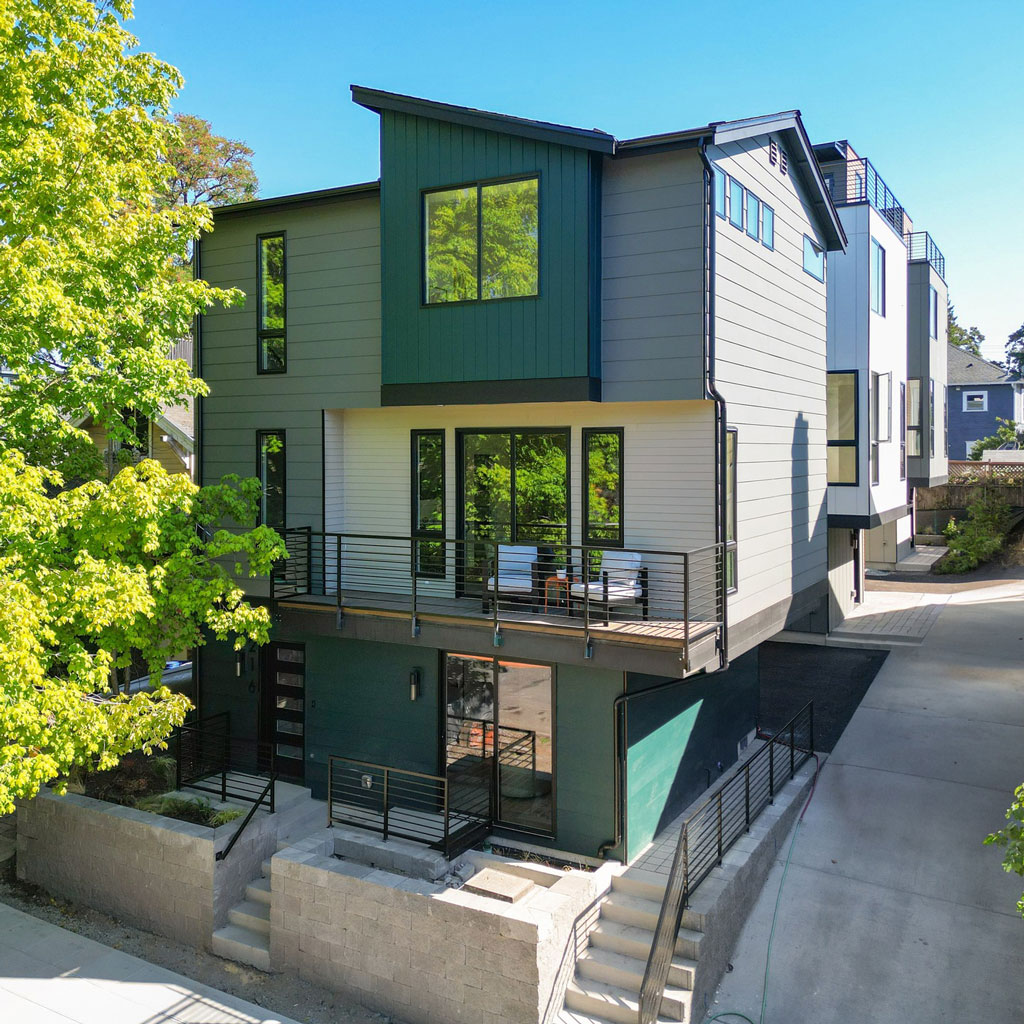 A modern multi-story townhouse with green and gray siding, a balcony, and large windows on a sunny day.
